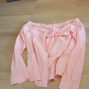 Ljusrosa offshoulder skjorta med knyt - Trendig ljusrosa skjorta i bomull med offshoulder-modell och långa ärmar. Skjortan har knappar framtill och en snygg knytning vid bysten som ger en cool och avslappnad vibe. Perfekt för dig som vill sticka ut lite extra med din stil.
