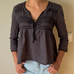 Odd molly blus - Supersöt mörkgrå blus från Odd Molly med trekvartsärm, v-ringning och knyt framtill. Blusen har broderade detaljer, små prickar i tyget och spetskant vid hals och ärmslut. Luftig och flowy passform, perfekt till jeans eller kjol.