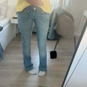 Wrangler ljusblå bootcut jeans - Säljer ett par ljusblå jeans från Wrangler med bootcut-snitt och klassiska fem fickor. Jeansen har låg midja och en stor Wrangler-logga på bakfickan. Perfekta för dig som gillar retrovibbar och vill ha ett par snygga jeans med lite utsvängda ben.