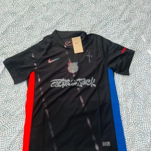 FC Barcelona x Nike Cactus Jack tröja - Snygg svart FC Barcelona fotbollströja från Nike i samarbete med Cactus Jack. Tröjan har korta ärmar, röda och blå sidopaneler, klubbmärke på bröstet och tryck med Cactus Jack-logga. Materialet är lätt och andas, perfekt för fotboll eller streetwear.