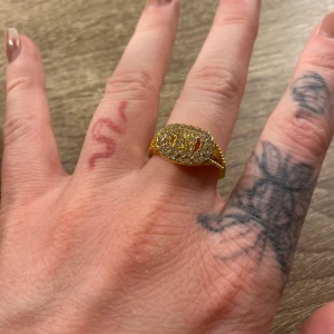 Guldig ring med glittrande stenar - Säljer en snygg guldig ring med oval topp täckt av glittrande stenar. Ringen har en chunky känsla och detaljerade sidor som ger extra bling. Perfekt för dig som vill sticka ut och addera lite extra shine till din stil.