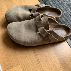 Beige mockasandaler med spänne - Snygga sandaler i beige mocka med rund tå och justerbart metallspänne. Skorna har en platt sula och öppen häl, vilket ger en avslappnad och trendig look. Perfekta för dig som gillar stilren och enkel design.
