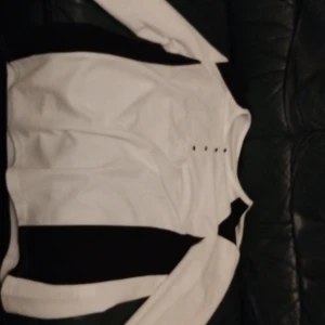 Vit och svart långärmad tröja med knappar som är väldigt snygg och passar väldigt bra in på de varmare dagarna på sommarn - Snygg långärmad tröja i vitt med svarta paneler längs sidorna och axlarna. Tröjan har rund halsringning och dekorativa knappar framtill. Perfekt för dig som gillar en stilren och sportig look.