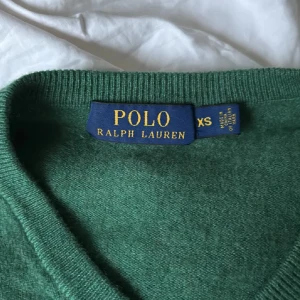 Grön merinoullströja Polo Ralph Lauren - Snygg grön tröja från Polo Ralph Lauren i 100% merinoull. V-ringad modell med lång ärm och klassisk broderad logga på bröstet. Perfekt för dig som gillar stilrena och tidlösa plagg med en lyxig känsla. Har några små hål men de går nog ganska enkelt att sy ihop! Annars bra skick och super mysig