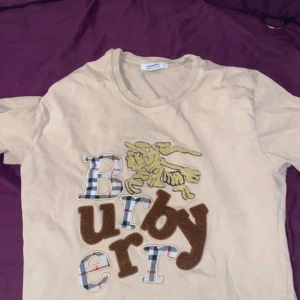 Beige Burberry t-shirt med brodyr - Snygg beige t-shirt från Burberry med coolt broderat Burberry-tryck framtill i rutigt och brunt samt en guldfärgad riddare. Klassisk rund hals och korta ärmar. Tillverkad i mjuk bomull, perfekt för en trendig och avslappnad stil.