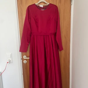 Långärmad vinröd maxiklänning - Vinröd långklänning från Jaani med rund halsringning och långa ärmar. Klänningen har en markerad midja och ett elegant, flytande fall. Perfekt för dig som vill sticka ut med en djup färg och klassisk siluett.