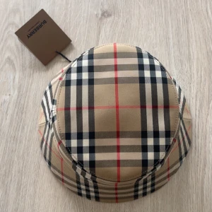 Burberry rutig bucket hat beige - Snygg bucket hat från Burberry i klassiskt beige rutigt mönster med svarta, vita och röda detaljer. Hatten har en mjuk form och är tillverkad i bomull. Perfekt accessoar för att lyfta din stil med en ikonisk touch. Nypris: 3300kr. Denna modell är liten i storlek, M är som S 
