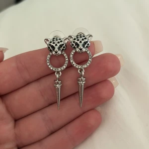 Silvriga leopardörhängen med strass - Coola örhängen i silverfärg med leopardhuvud i svart och vitt, glittrande strassringar och spetsiga hängen. Perfekt statement-accessoar för dig som vill sticka ut med djurmönster och edgy detaljer.
