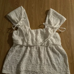 Vit smockad linnetopp med knytband - Supersöt vit linnetopp med smockad struktur och breda axelband som knyts i rosetter. Toppen är ärmlös och har en luftig, lätt utställd passform. Perfekt för varma dagar och ger en romantisk vibe till din outfit.