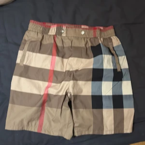 Burberry rutiga shorts i beige och rött - Snygga Burberry shorts med klassiskt rutigt mönster i beige, brunt, svart, vitt och rött. De har elastisk midja med knappar och dragsko, samt fickor på sidorna och bak. Perfekta för sommaren och ger en lyxig vibe till din outfit.
