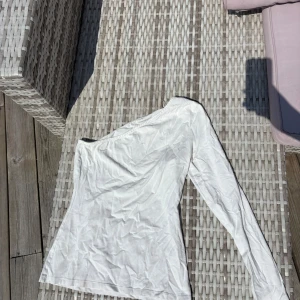 Vit oneshoulder topp från NELLY - Säljer en stilren vit oneshoulder topp från NELLY Trend i storlek S. Toppen har lång ärm på ena sidan och är axelbandslös på andra, vilket ger en cool asymmetrisk look. 