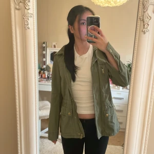 Olivgrön vindjacka med dragkedja och knappar - Snygg olivgrön vindjacka med hög krage, dragkedja och knappar framtill. Jackan har flera fickor med guldiga detaljer och justerbar midja. Perfekt för lager-på-lager och enkel att matcha till olika outfits.