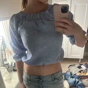 Blåvit randig croppad blus med knyt i ryggen - Säljer en blåvit randig croppad blus med resår vid halsringningen och knytning i ryggen. Blusen har trekvartsärmar och är perfekt för varmare dagar. Snygg och trendig modell som passar till jeans eller kjol.