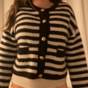 Svartvit randig kofta med guldknappar - Säljer en fluffig svartvit randig kofta med guldknappar framtill och två små fickor. Koftan är croppad och har långa ärmar. Perfekt för dig som gillar en trendig stil! Använd men inga defekter 💞Nypris 600kr 