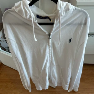Vit hoodie från Polo Ralph Lauren - Säljer en klassisk vit hoodie från Polo Ralph Lauren med dragkedja och huva. Tröjan har två fickor framtill och den ikoniska broderade loggan i mörkblått på bröstet. Perfekt för en avslappnad stil.