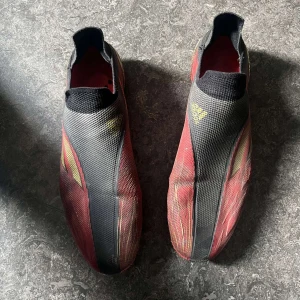 Adidas X Speedflow + fotbollskor - Säljer ett par Adidas fotbollsskor i rött och svart med guldiga detaljer. Skorna har en strömlinjeformad design utan snörning och mönstrad ovandel. Sulan har fasta dobbar för spel på gräs. Perfekta för dig som vill sticka ut på planen. 