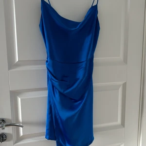Blå satin klänning från Zara - Säljer en blå klänning från Zara i satin med smala axelband och draperad detalj framtill. Klänningen har en elegant, figurnära passform och är perfekt för sommarens tillfällen.