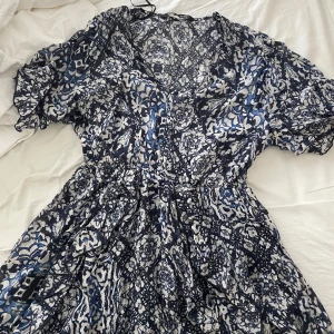 Blommig blå och vit klänning från ZARA - Säljer en blommig klänning i blått och vitt med v-ringning och korta ärmar. Klänningen har en markerad midja och ett luftigt fall, perfekt för varma dagar. Mönstret är detaljerat och ger en bohemisk känsla. Köpt för 399 säljs nu för 100