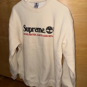 Vit sweatshirt från Supreme x Timberland - Vit sweatshirt från Supreme och Timberland med broderad logga och texten 'WIND, WATER, EARTH AND SKY.' på bröstet. Tröjan har rund hals och långa ärmar. Tillverkad i 100% bomull. Passar Herr Large.