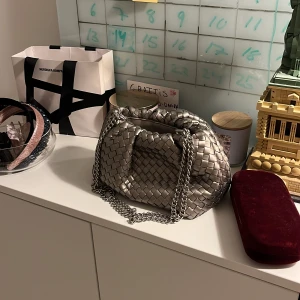 Silver axelväska med flätad design - Säljer en snygg silverfärgad axelväska med flätad yta och kedjerem. Väskan har en mjuk, rundad form. Väskan är från Gina och endast använd en gång. 