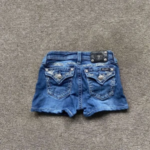 Blå jeansshorts från missme - Snygga blå jeansshorts från missme med dekorativa detaljer på fickorna. låg midja och kort passform. Midjemått tvärsöver 35cm och total längd 27cm