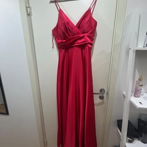Röd långklänning med omlott och smala axelband - Säljer en elegant röd långklänning med omlott framtill och smala axelband. Klänningen har en markerad midja och ett mjukt, glansigt tyg som faller snyggt. Perfekt för bal eller festliga tillfällen.