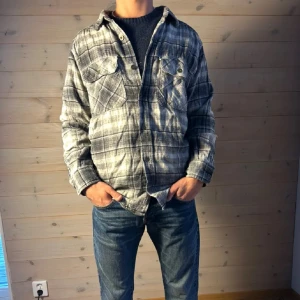 Flanellskjorta  - Säljer en grå och svart rutig skjorta med knappar och två bröstfickor. Skjortan har lång ärm och klassisk krage, perfekt för en avslappnad stil. Passar dig som gillar en normal passform och vill ha något stilrent till vardags.