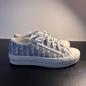 Gråvita sneakers från Christian Dior - Säljer ett par snygga sneakers från Christian Dior i grått och vitt med monogrammönster. Skorna har vit sula, rund tå och snörning framtill. Märkeslogga på tungan och på snörningen. Perfekta för dig som gillar exklusiva detaljer.