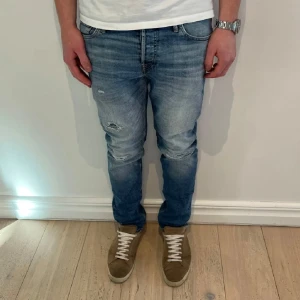 Jack and Jones Blå slitna jeans med raka ben - Säljer ett par blå jeans med slitningar och ljusa partier på låren och knäna. Jeansen har klassisk femficksdesign, dragkedja och knapp i midjan. Passformen är straight och materialet är denim. Sittter som slim fit 