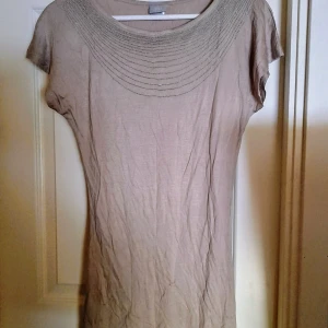 Beige kortärmad topp blus med dekorativ halsringning - Säljer en enkel och stilren beige t-shirt med korta ärmar och rund halsringning med dekorativa sömmar. Den har en rak passform och är tillverkad i ett mjukt, tunt material. Sparsamt använd. Storlek xs