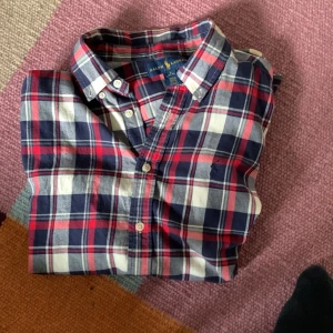 Rutig skjorta från Ralph Lauren - Klassisk rutig skjorta från Ralph Lauren i blått, rött och vitt. Skjortan har knappar framtill, lång ärm och den ikoniska broderade loggan på bröstet. Perfekt för en stilren och avslappnad look.