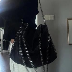 Svart axelväska med kedjedetaljer - Säljer denna inspirerade svarta tiamo axelväskan med snygga silverfärgade kedjor längs kanterna och som axelrem. Väskan är rymlig och passar superbra till typ allt!💕