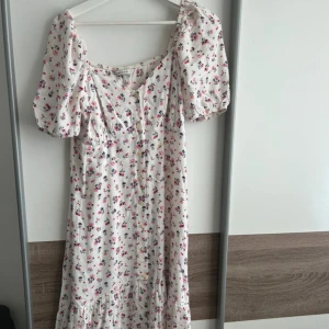 Vit blommig långklänning med puffärmar - Romantisk långklänning i vit bas med små blommor i rosa och blått. Klänningen har puffiga kortärmar, v-ringning och knappar framtill. Volangdetalj nedtill ger extra rörelse och sommarkänsla. Perfekt för varma dagar och fina tillfällen.