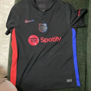FC Barcelona svart fotbollströja Nike - Säljer en svart FC Barcelona fotbollströja från Nike med röda och blå detaljer på sidorna. Tröjan har korta ärmar, klubbmärke på bröstet, Spotify-logga framtill och UNHCR-logga bak. Tillverkad i Dri-FIT-material för bra komfort.