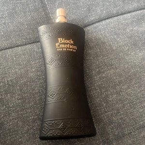 Black Emotion Eau de Parfum - Säljer en parfymflaska Black Emotion Eau de Parfum, 100 ml. Flaskan är matt svart med dekorativa mönster och har en elegant, kurvig form. Texten på flaskan är i guld. Perfekt för dig som gillar unika och stilrena doftflaskor.