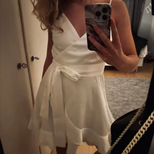 Vit klänning  - Säljer en söt vit omlottklänning med knytband i midjan och v-ringad halsringning. Klänningen är ärmlös och har en kort, svängd kjol som ger en lätt och somrig känsla. Perfekt för varma dagar eller speciella tillfällen.