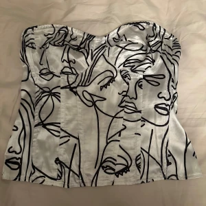 Vit korsettopp med svart ansiktsmönster - Säljer en snygg vit korsettopp med svart abstrakt ansiktsmönster. Toppen har en markerad byst och dragkedja bak. Perfekt för dig som vill sticka ut med en unik och trendig look.