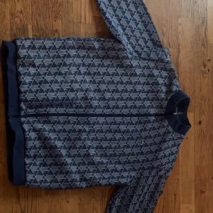 Mönstrad track jacket från Lacoste - Säljer en blå och grå track jacket från Lacoste med hel dragkedja och ribbade muddar. Jackan har ett grafiskt mönster över hela ytan och den klassiska krokodilloggan på bröstet. Perfekt för dig som gillar sportig och stilren look.