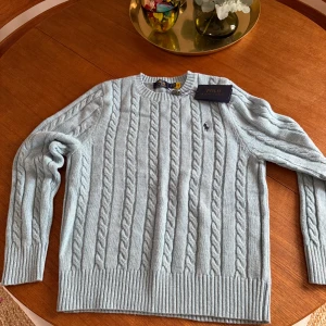 Ljusblå kabelstickad tröja från Polo Ralph Lauren - Säljer en ljusblå kabelstickad tröja från Polo Ralph Lauren. Tröjan har rund halsringning, ribbade muddar och det klassiska broderade logotypen på bröstet. Perfekt för sena sommar kvällar. Skick 10/10