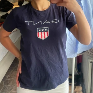 Mörkblå t-shirt från Gant - Säljer en mörkblå t-shirt från Gant med klassisk logga och sköld med stjärnor och ränder på bröstet. T-shirten har rund halsringning och korta ärmar. Perfekt för en avslappnad och stilren look.