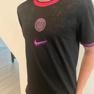 PSG x Jordan svart fotbollströja - Snygg svart PSG fotbollströja från Nike med rosa och lila detaljer. Tröjan har korta ärmar, rund halsringning och diskret blommönster i tyget. PSG-logga och Nike swoosh i rosa framtill. Perfekt för dig som gillar stilrena matchtröjor.