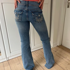 Blå bootcut jeans med broderade bakfickor - Säljer ett par blå bootcut jeans från H&M med snygga broderade detaljer på bakfickorna och klassisk femficksdesign. Jeansen har låg midja och är tillverkade i denim med lätt stretch. Perfekta för dig som gillar en retroinspirerad look.