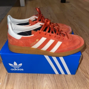 Adidas Handball Spezial red/orange sneakers - Säljer ett par Adidas Spezial sneakers i röd/orange mocka med vita detaljer och klassiska tre ränder på sidan. Använda 2-3 ggr, perfekt skick förutom lite ljusare i färgen vid tårna. Orginalpris 1299kr. 