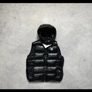 Bra väst inför sommaren - Snygg svart dunväst från Moncler med glansig finish och huva. Västen har en klassisk puffer-design, dragkedja framtill och Moncler-logga på bröstet. Perfekt för lager-på-lager och håller dig varm under kyliga dagar. Pris kan sänkas vid snabb affär. Intresserad vid byten också