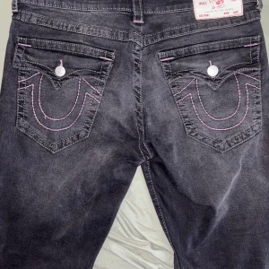 Svarta jeansbyxor från True Religion med rosa / lila sömmar - Säljer ett par svarta jeansbyxor från True Religion, modell Geno Relaxed Slim. Byxorna har rosa / lila kontrastsömmar och klassiska fickdetaljer med knappar. Snygg passform och unika bakfickor med broderad logga. W33 L33 Skriv om du har frågor!