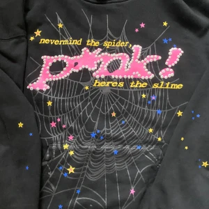 Svart hoodie med tryck från SP5DER - Cool svart hoodie från SP5R med tryck i rosa, vitt och gult framtill. Texten 'never mind the spider, heres the slime' och ett stort 'prak!' i pärl-liknande stil. Spindelnätsmönster och stjärnor över hela framsidan. Perfekt statement-plagg!