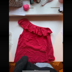 Röd one shoulder topp med volang - Säljer en snygg röd one shoulder topp med volangdetalj längs halsringningen. Perfekt för dig som vill sticka ut med en trendig och färgstark look. Tunn och lätt i materialet, passar bra till sommar dagar.