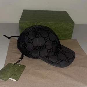 Svart keps från Gucci med GG-mönster - Snygg svart keps från Gucci med klassiskt GG-mönster över hela kepsen. Justerbar rem baktill i skinn och detaljer i guld. Kepsen är i bomullsmaterial och har märkets logga på etiketten.