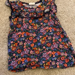 Blommig ärmlös topp från H&M - Färgglad ärmlös topp från H&M med blommigt mönster i rött, lila, blått och orange på mörk botten. Toppen har volangdetaljer vid axlarna och halsringningen. Perfekt för att liva upp garderoben med lite färg!