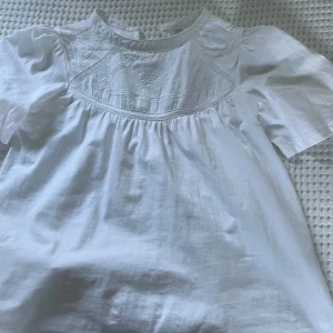 Vit blus med broderad detalj från Zara - Säljer en söt vit blus från Zara med korta ärmar och broderade detaljer upptill. Säljer då den är för liten i storlek och inte kommit till användning. Perfekt nu till våren och sommaren!